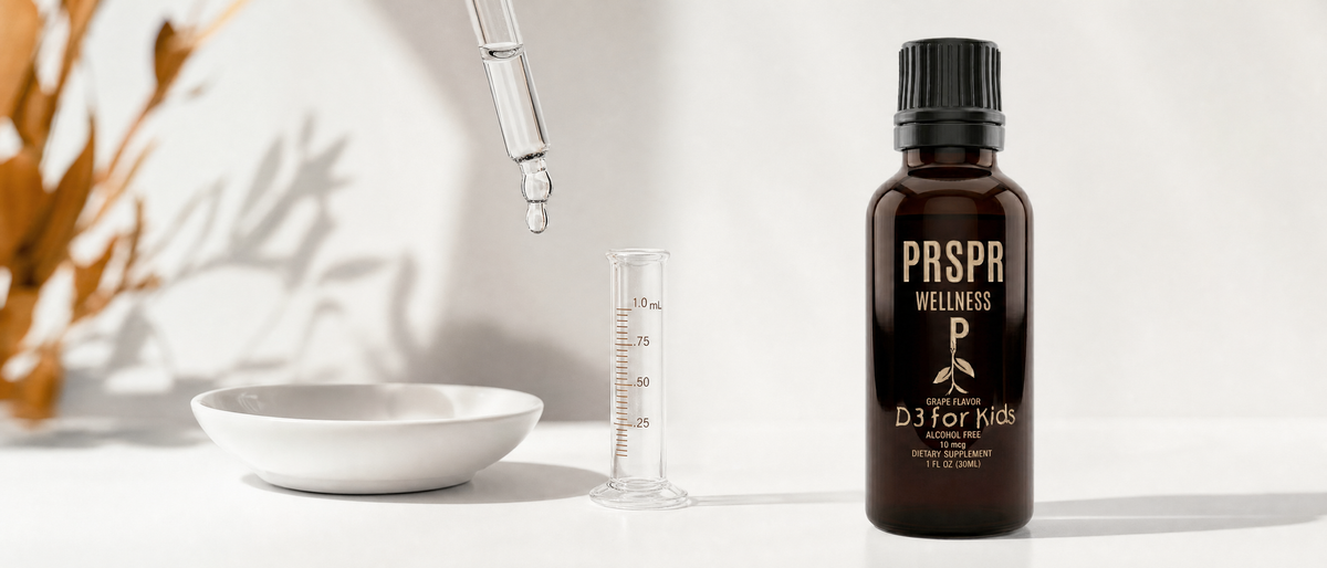 PRSPR Wellness Children's Liquid Vitamin D3 bottle on white beside an upright glass pipette forming a droplet and a measuring cylinder