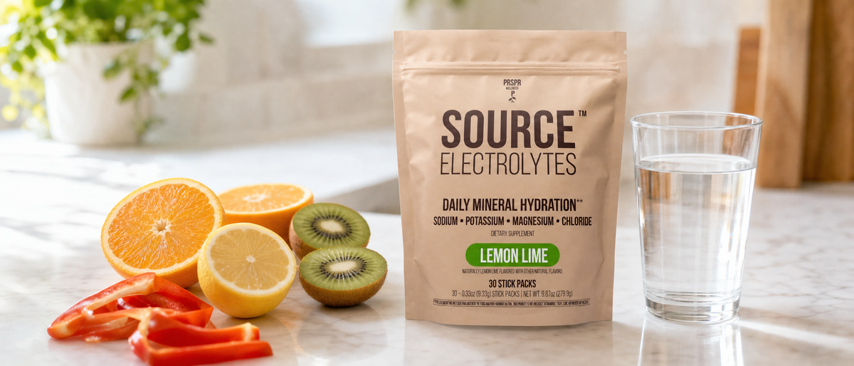 Source Electrolytes Lemon Lime pouch on a white marble counter beside halved oranges, a lemon, kiwi, red bell pepper strips and a glass of water