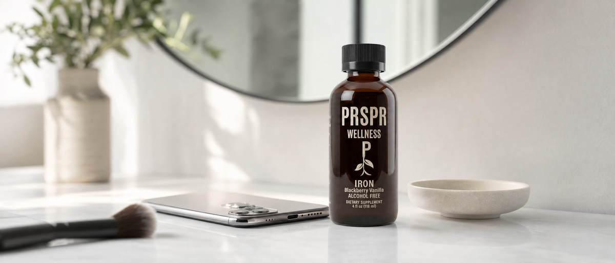PRSPR Liquid Iron bottle on a white bathroom vanity in front of a round mirror, beside a phone lying face down and a makeup brush