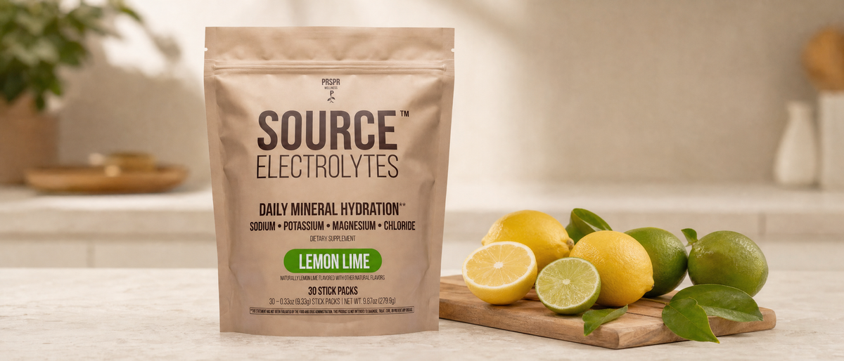 A PRSPR Wellness Source Electrolytes Lemon Lime pouch on a stone counter beside whole and halved lemons and limes on a wooden board