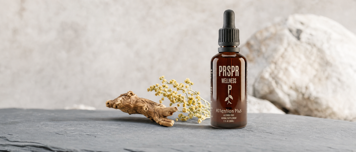 PRSPR Wellness Attention Plus dropper bottle on a slate surface beside a dried root and small yellow flowers