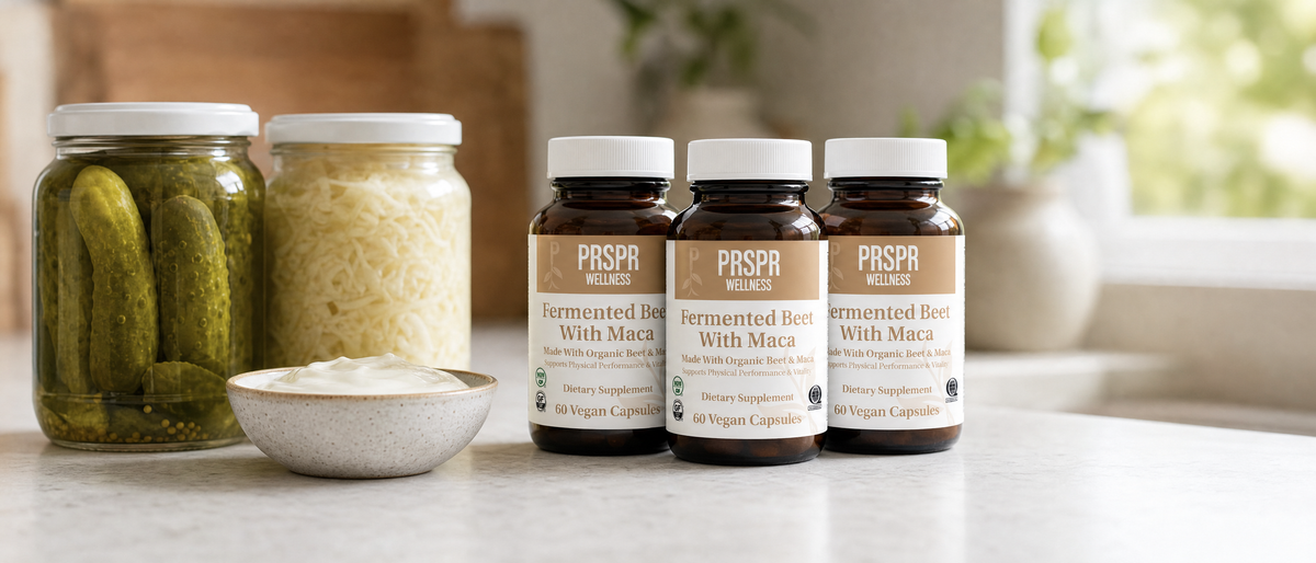 Three PRSPR Wellness Fermented Beet with Maca bottles beside jars of pickles and sauerkraut and a bowl of yogurt
