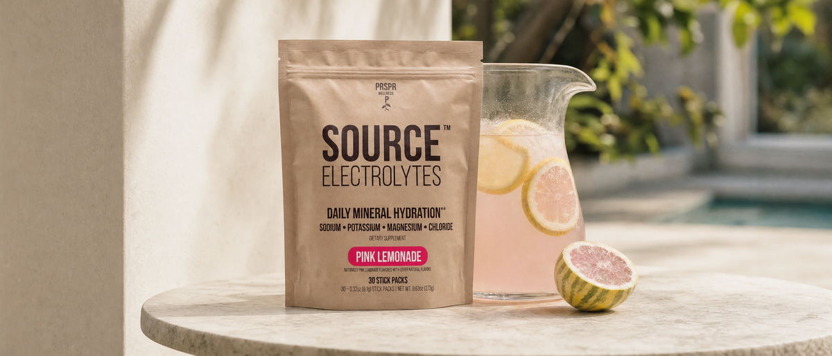Source Electrolytes Pink Lemonade pouch on a marble table beside a pitcher of pink lemonade and a halved variegated pink lemon