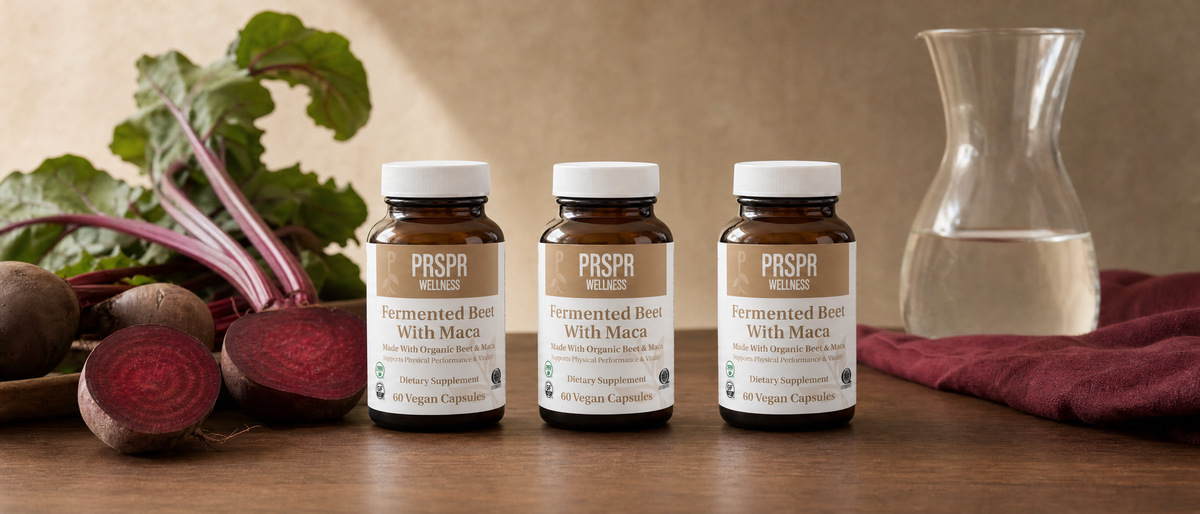 Three jars of PRSPR Wellness Fermented Beet With Maca on a walnut table beside fresh beetroots, a halved beet, a red linen napkin and a glass carafe of water