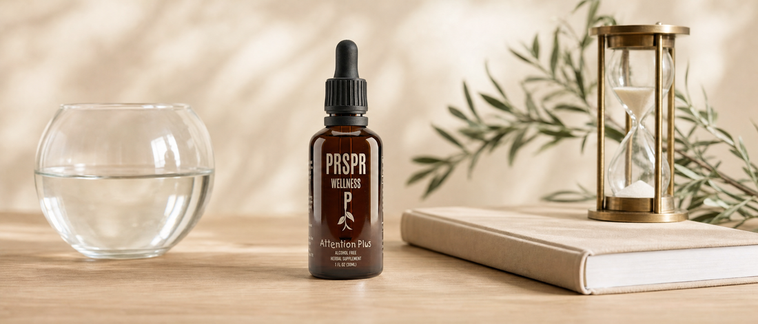 A PRSPR Wellness Attention Plus dropper bottle on a wooden desk beside an empty glass fishbowl of water, a closed notebook and a brass hourglass