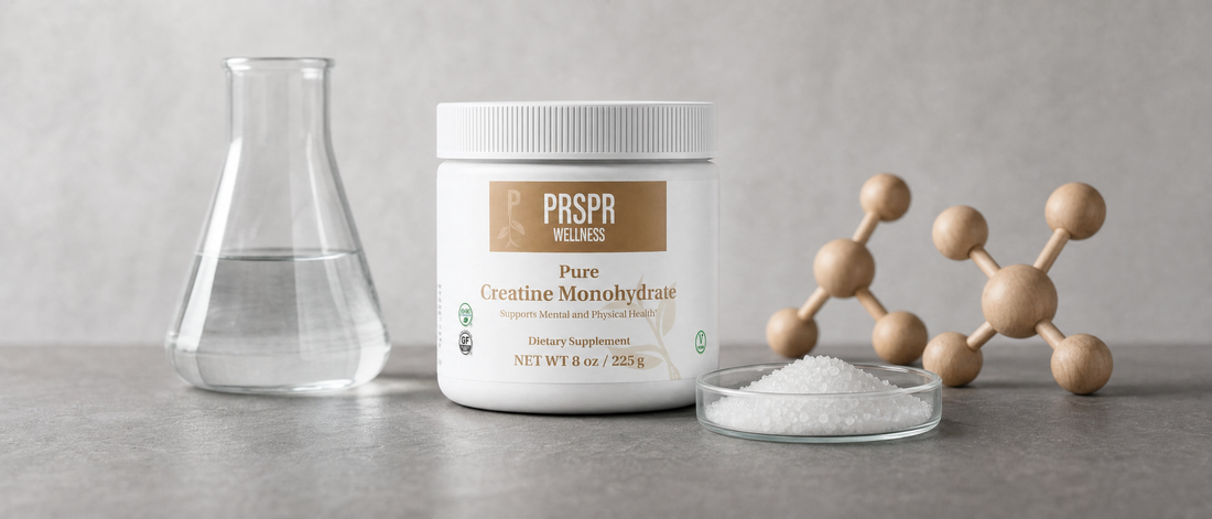 A white tub of PRSPR Pure Creatine Monohydrate on grey slate beside a glass flask of water, a small dish of white crystals and a wooden ball-and-stick molecular model.