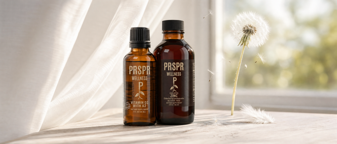 Two PRSPR Wellness amber bottles, Vitamin D3 with K2 and Chelated Zinc, on a whitewashed table by a window with a sheer curtain, a dandelion seed head and white feathers.